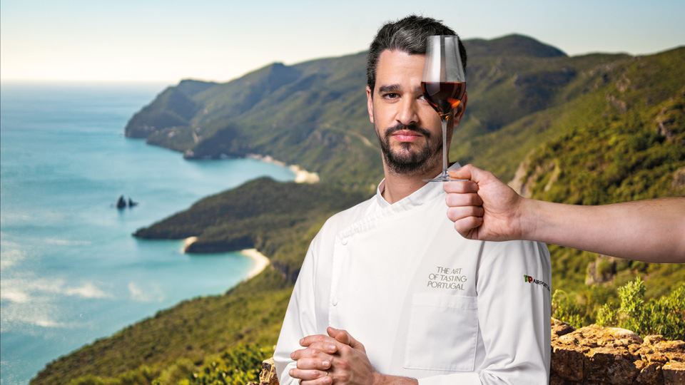 Medium shot of Chef André Cruz, dressed in a white chef's coat, with his arms slumped, half-bent below his chest, the fingers of both hands intertwined. On the right side, there is an arm holding a glass of Moscatel de Setúbal, which is superimposed on the left side of the chef's face. In the background is a panoramic view of the green mountains of the Setúbal Peninsula. 
