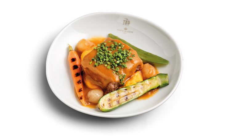 Photography of a deep white plate with the golden TAP Air Portugal logo on the rim, containing a piece of veal Osso Buco in the center, surrounded by orange Moscatel de Setúbal sauce, carrot puree and spring vegetables.