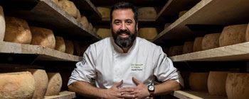 Full body photograph of Chef Cláudio Pontes. The Chef wears a kitchen coat on the left side of which, on the chest, you can read the embroidered inscription "Da origem à terra" (From the origin to the land), as well as his name. The Chef is sitting with his arms crossed, in the middle of two wooden shelves where several large cheeses are being matured.