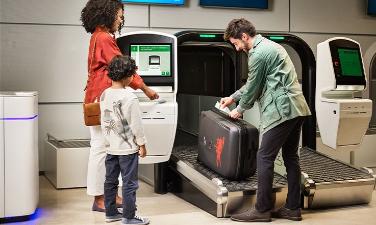 Imagem de uma jovem e sorridente família no aeroporto que, na área de self-check-in, se prepara para etiquetar e despachar a sua bagagem de porão.