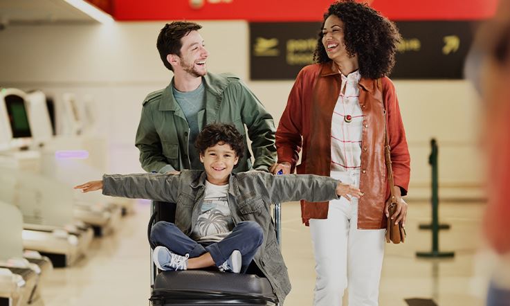 Fotografia de uma alegre família que caminha no aeroporto com um carrinho de bagagem. Sobre o carrinho de bagagem encontra-se um jovem, sentado, de braços abertos e com uma expressão sorridente. O ambiente no aeroporto é calmo e luminoso.