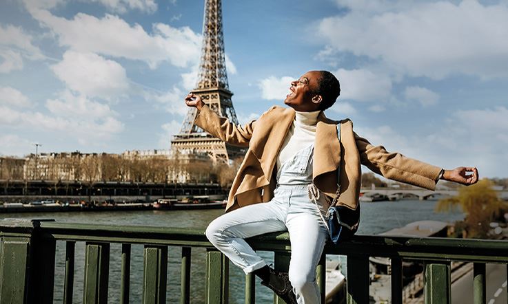 Fotografia de uma jovem mulher sorridente envergando roupas claras e elegantes. A jovem está sentada em cima do varão de uma ponte, de braços abertos e estendidos, e tem atrás de si uma paisagem típica parisiense, onde se vê o rio Sena e a torre Eiffel.