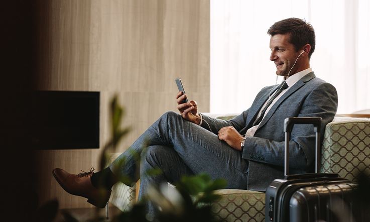 Fotografia de corpo inteiro de um homem envergando fato e gravata. Está sentado numa cadeira de aspecto confortável, numa sala, e segura um smartphone com a mão direita, para cujo ecrã olha sorridente.