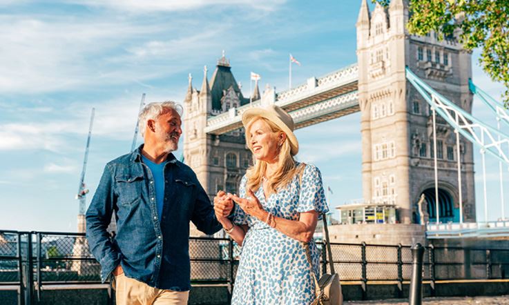 Fotografia de um casal envergando roupa de meia estação num dia muito luminoso. Atrás do casal, é visível em segundo plano a conhecida Tower Bridge de Londres.