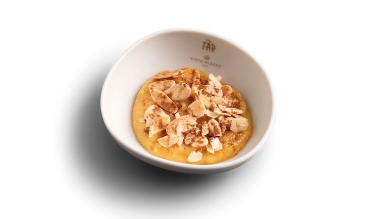 Photograph of a deep, white plate, with the golden TAP Air Portugal logo on the edge, containing a generous portion of the golden soup dessert, sprinkled with a layer of sliced almonds and cinnamon.