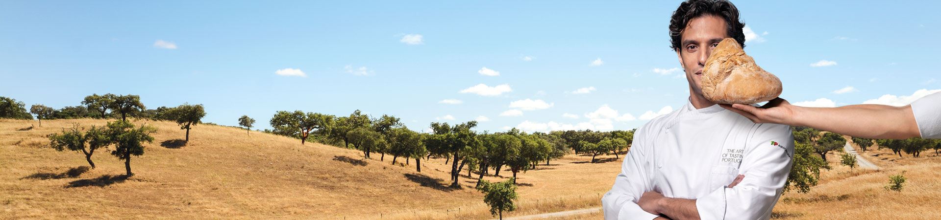 Średnie zdjęcie szefa kuchni Carlosa Afonso. Ubrany jest w biały fartuch kuchenny z napisem „The art of tasting Portugal” na lewej górnej kieszeni nad klatką piersiową. Na lewym rękawie widoczne jest logo TAP Air Portugal. Za szefem kuchni, w tle, znajduje się panoramiczne zdjęcie równin Alentejo z suchą, złotą glebą, usianą kilkoma rozrzuconymi dębami ostrolistnymi i/lub dębami korkowymi. Po prawej stronie obrazu pojawia się ręka trzymająca kilogram chleba Alentejo przed szefem kuchni, częściowo zakrywająca lewą stronę jego twarzy. 