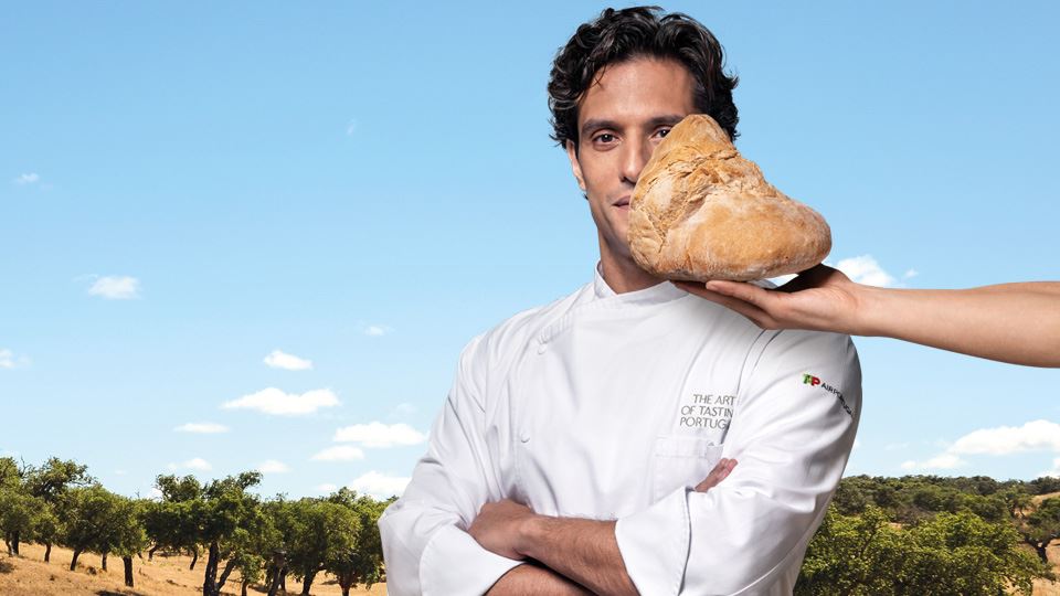 Medium shot photograph of Chef Carlos Afonso. He is dressed in a white kitchen coat that has "The art of tasting Portugal" written on the upper left pocket, over the chest. The TAP Air Portugal logo can be seen on the left arm sleeve. Behind the Chef, in the background, there is a panoramic photograph of the plains of Alentejo with dry, golden soil, dotted with a few scattered holm oaks and/or cork oaks. On the right side of the image, an arm appears holding a kilo of Alentejo bread in front of the Chef, partially covering the left side of his face.