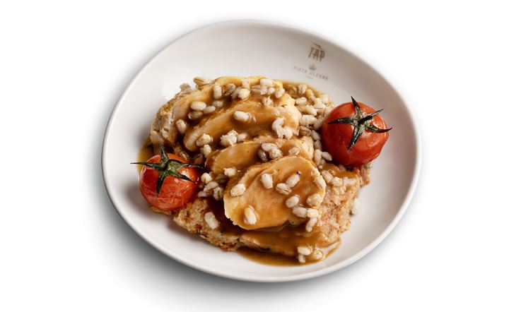 Photograph of a shallow white plate, with the golden TAP Air Portugal logo on the rim, inside which is a bed of tomato crumbs and Alentejo bread, topped by four slices of chicken breast, flanked on the right and left by a small roasted cherry tomato. On top of these ingredients, a few grains of barley are scattered alongside a generous layer of sauce.