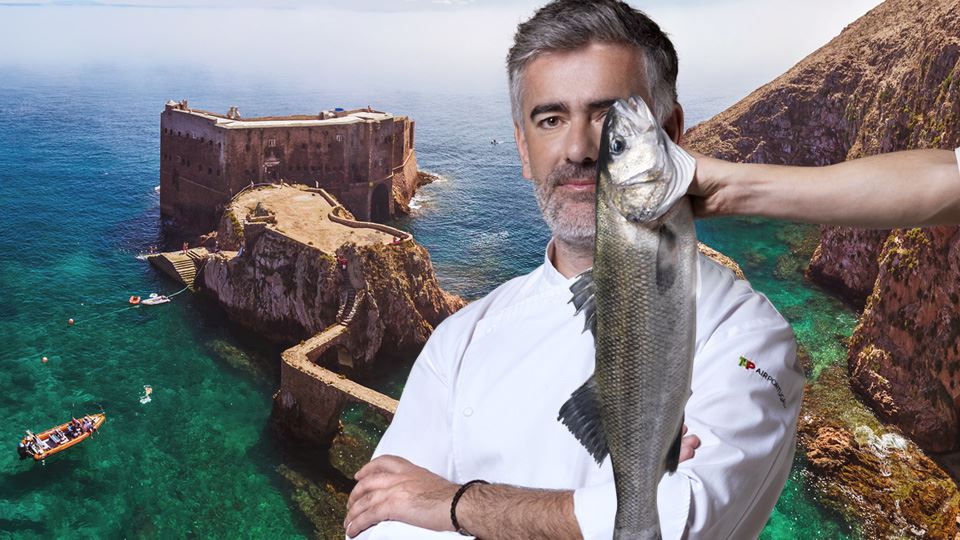 Photographie en plan moyen du chef Alexandre Silva. Il porte une veste de cuisine blanche avec le logo TAP Air Portugal brodé sur le bras gauche. Derrière le chef, en arrière-plan, une photographie panoramique de la mer et de la falaise, au centre de laquelle se détache l'impressionnant Forte de São João Baptista, situé sur l'île de Berlenga Grande. Sur le côté droit de l'image, un bras tient un bar de Peniche par ses branchies, couvrant partiellement le côté gauche du visage du chef.