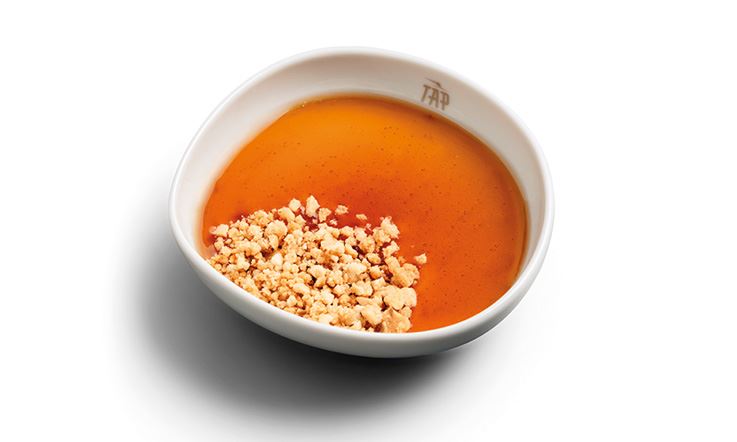Photographie d'une assiette creuse blanche dont le bord est marqué du logo doré TAP Air Portugal, contenant du pudding au lait de brebis recouvert d'un liquide orange et saupoudré de crumble beige, qui recouvre environ 1/3 du pudding.