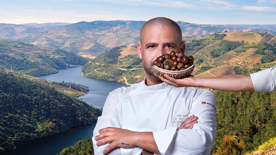 Medium shot photograph of Chef Oscar Geadas. He is dressed in a white kitchen coat that has "The art of tasting Portugal" written on the upper left pocket, on the chest, and the TAP Air Portugal logo on the left arm. Behind the Chef, in the background, there is a panoramic photograph of a region in northern Portugal.