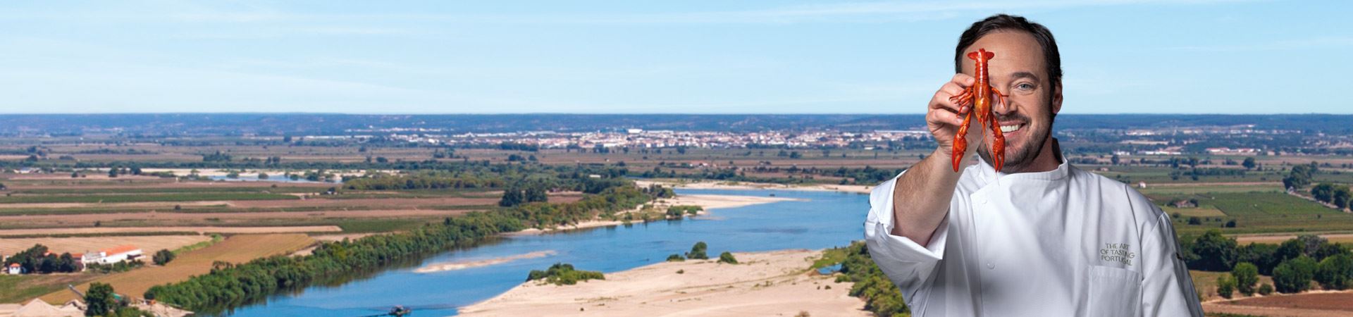 Panoramic photograph of a landscape in the Lezíria do Tejo region, showing a river, with sand and vegetation around it, a plain and vegetation, and in the background a village. On the right-hand side, above the photograph, is Chef Rodrigo Castelo, who holds a river crayfish in his left hand, which partially covers the right half of his face.