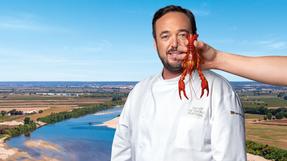 Panoramic photograph of a landscape in the Lezíria do Tejo region, showing a river, with sand and vegetation around it, a plain and vegetation, and in the background a village. On the right-hand side, above the photograph, is Chef Rodrigo Castelo, whose face is partially covered by a river crayfish.